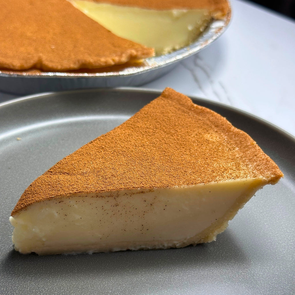 Slice of milktart on a gray plate with the rest of the milktart in the background.